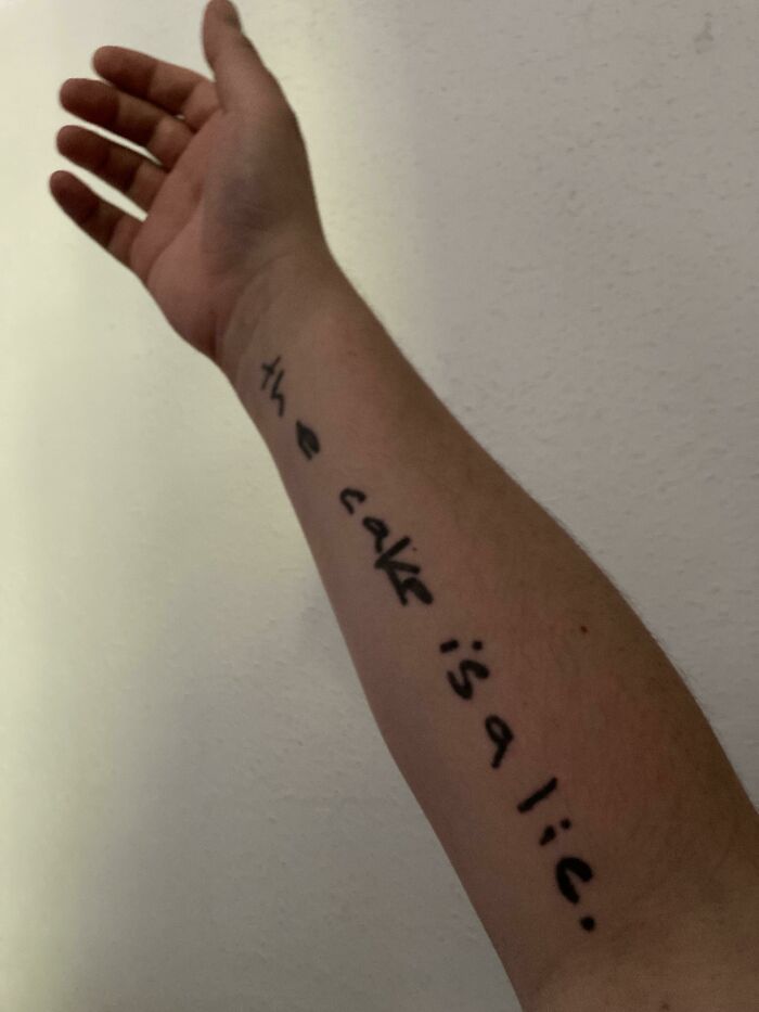 Arm tattoo text saying "the cake is a lie," highlighting tattoo artist choice.