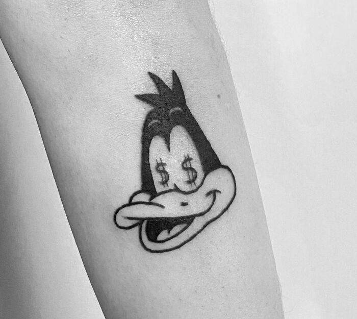 Cartoon tattoo of a duck character on an arm, eyes replaced with dollar signs, inspired by animated shows.