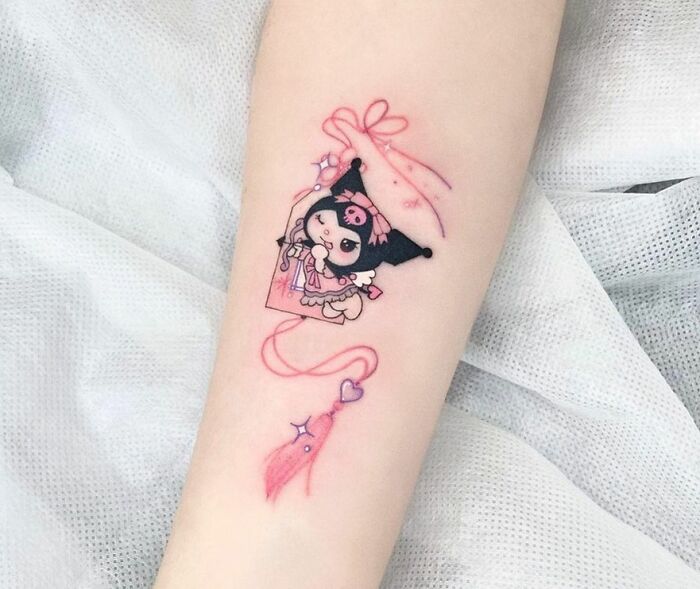Cute cartoon tattoo Kuromi Omamori with pink ribbon and hearts on forearm.