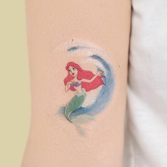 Cartoon tattoo of a mermaid Ariel with red hair and green tail in a wave design on forearm.
