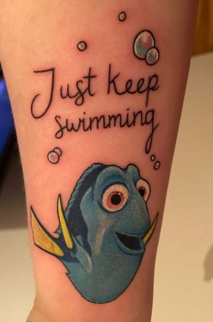 Cartoon tattoo of a blue fish Dory with yellow fins, with the phrase "Just keep swimming" above, surrounded by bubbles.
