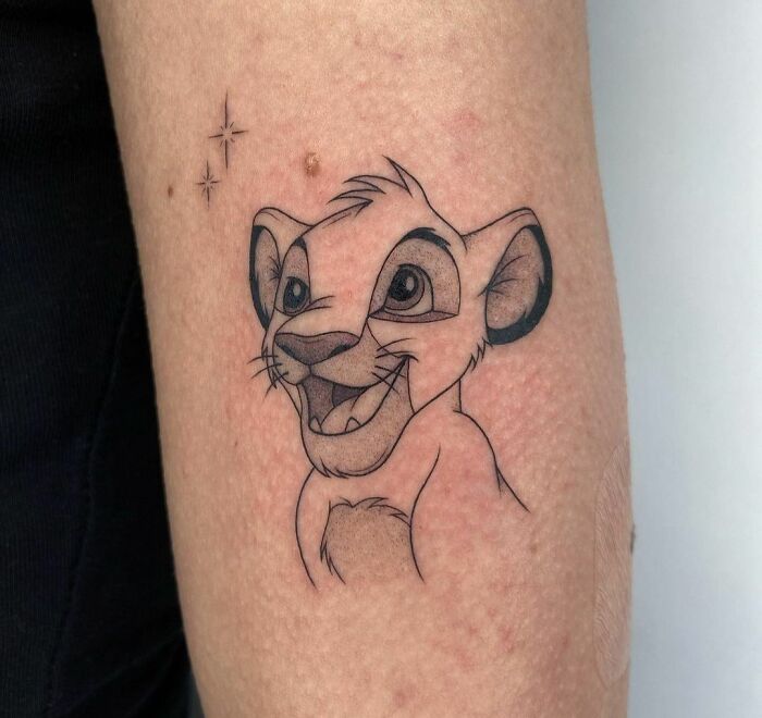 Cartoon tattoo of a smiling lion Simba cub on the forearm, evoking nostalgia with playful stars above.