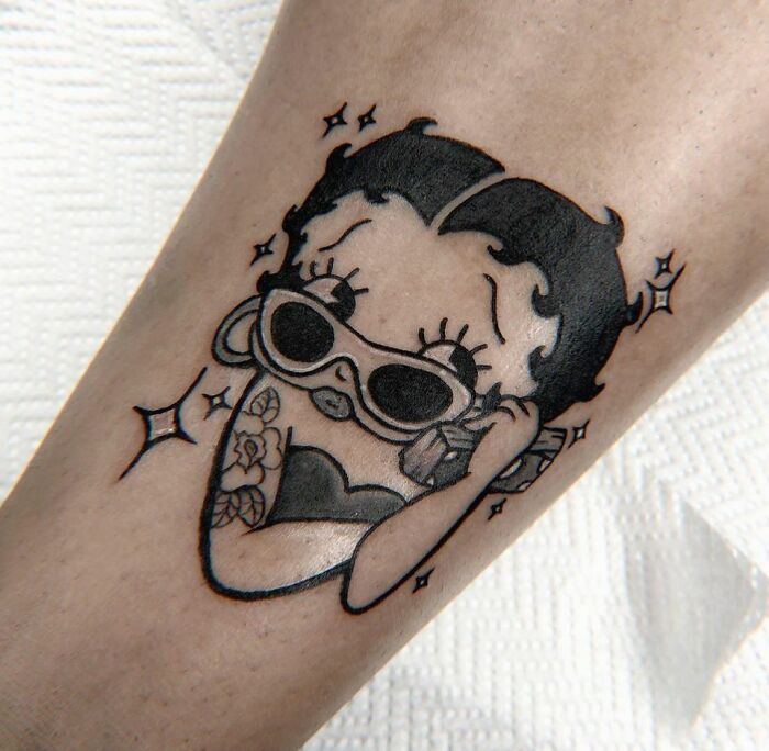 Cartoon tattoo featuring a character Betty Boop with sunglasses and floral arm design, embodying nostalgic style.