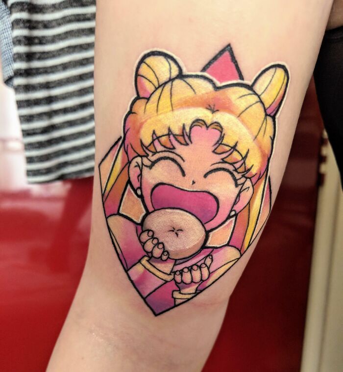 Cartoon tattoo of an ecstatic Sailor Moon character eating a donut on forearm, showcasing vibrant colors and nostalgic design.