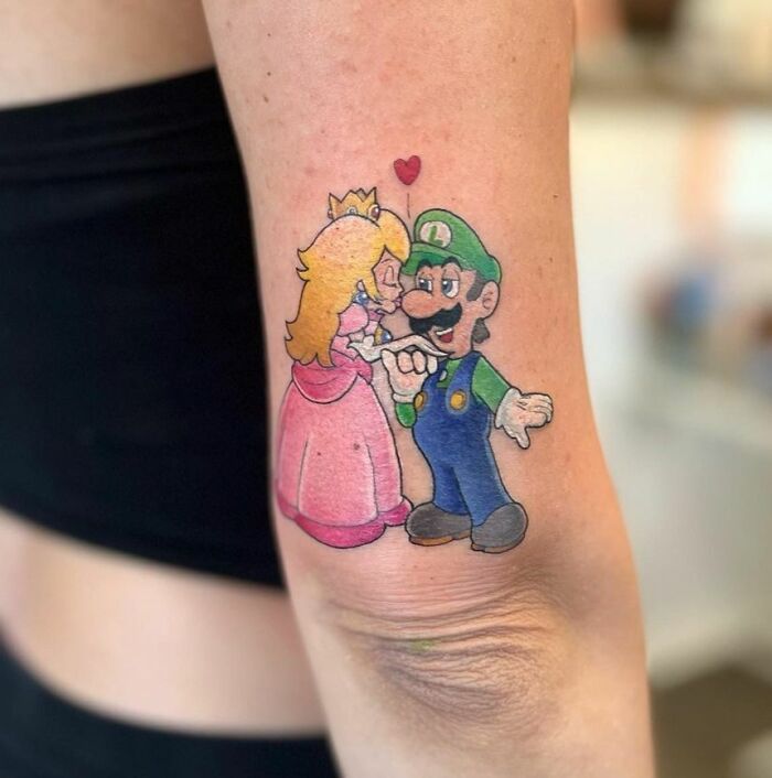 Cartoon tattoo of a princess Peach and plumber Luigi character, with a heart above, on a person's arm.