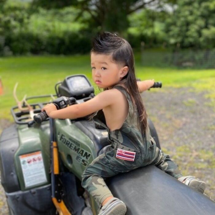 Young boy with a Cheddar Wiz mullet hairstyle sitting on an ATV outdoors in a casual, green setting.