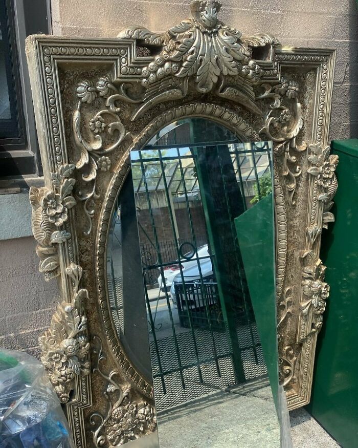 Ornate vintage mirror with intricate floral carvings, discarded on a New York City sidewalk among trash bins.