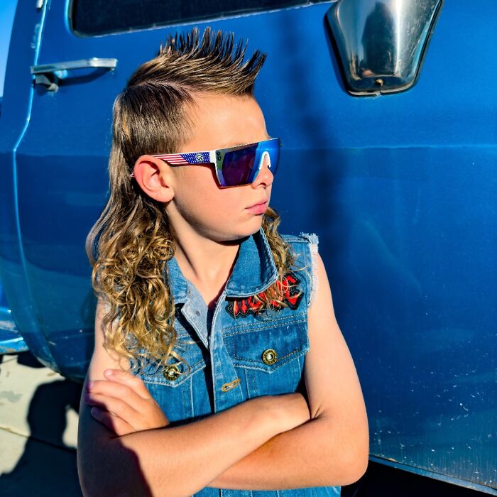 Boy with a standout cheddar wiz mullet hairstyle wearing patriotic sunglasses and a denim vest by a blue vehicle.