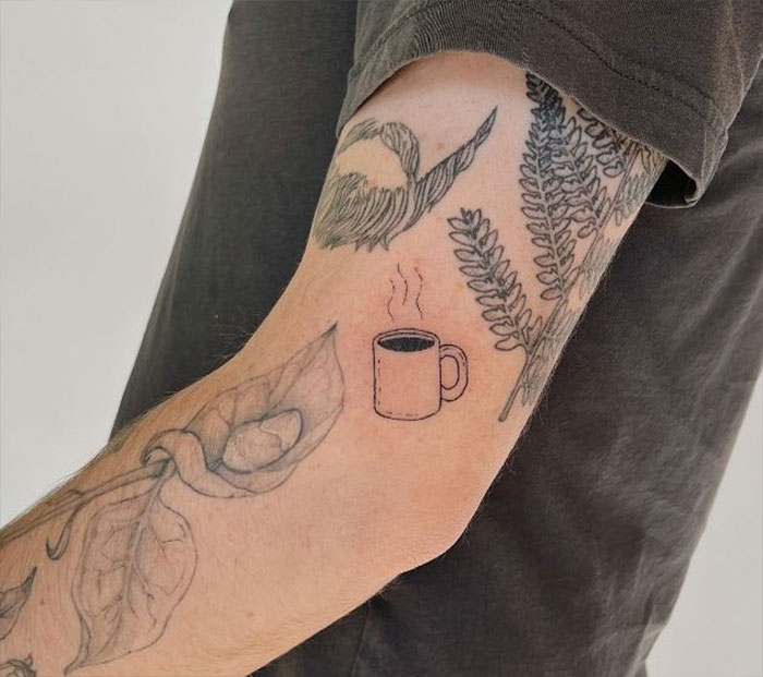 Elbow tattoo with coffee cup, bird, and fern designs on a person's arm wearing a dark shirt.