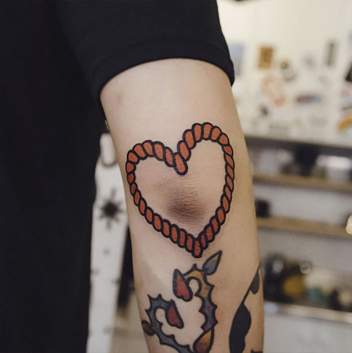 Heart-shaped design elbow tattoo with red rope outline on an arm, showcasing unique tattoo ideas.