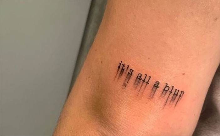 Elbow tattoo with text "it's all a blur" in a blurred style, on a person's arm.