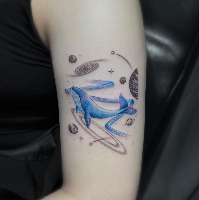 Cool space tattoo featuring a blue whale among planets and galaxies, artistically inked on a person's arm.