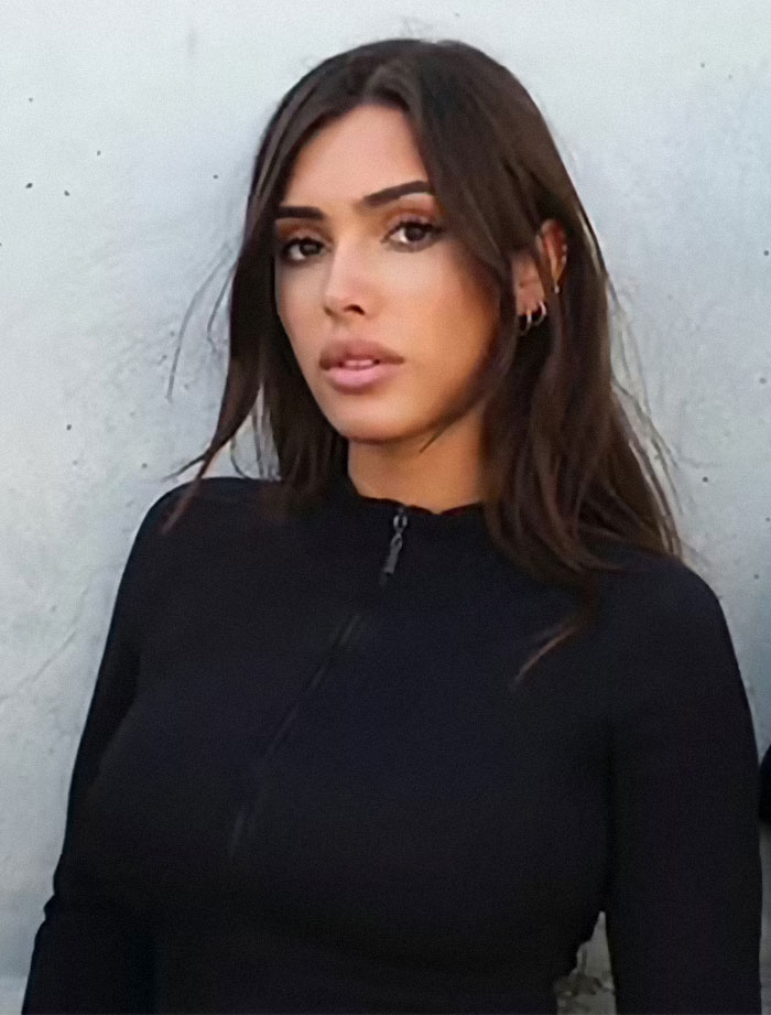 Bianca Censori in a black outfit, standing against a wall, with minimalistic style.