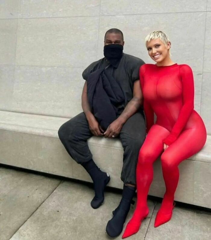 Bianca Censori in a revealing red outfit sitting on a bench next to Kanye West.