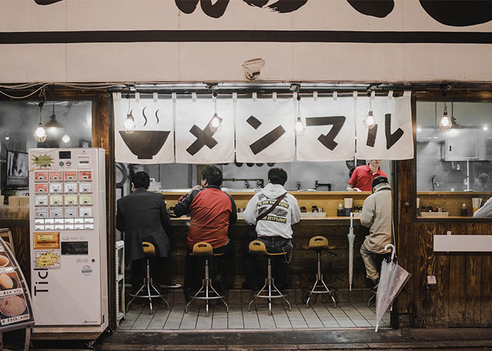 Customers seated at a small restaurant counter under hanging lights showing restaurant green flags to eat there.