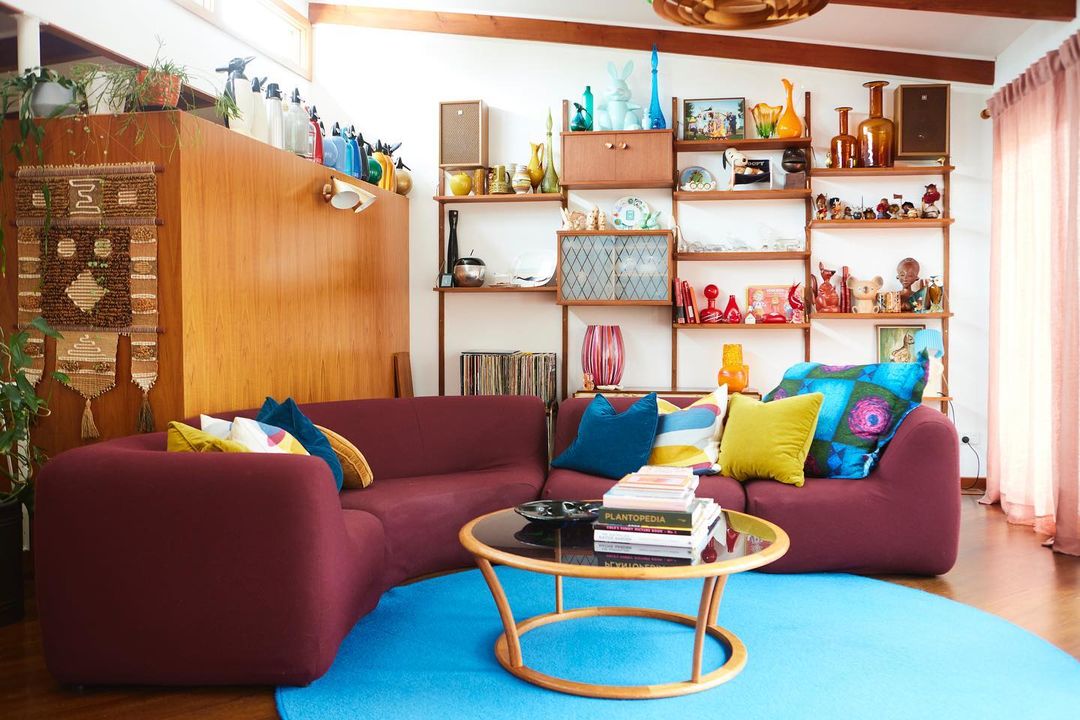 Bright living room with a large couch, colorful pillows, and a wooden shelf showcasing decorative items and books.