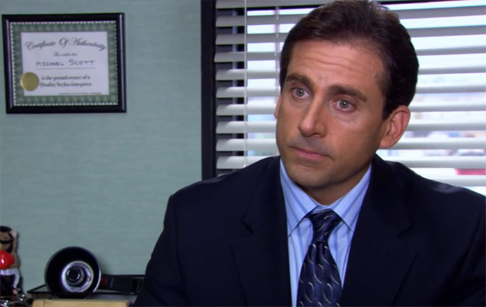 Michael Scott in a dark suit and tie, sitting in an office with blinds and a certificate, exemplifying best boss quotes.