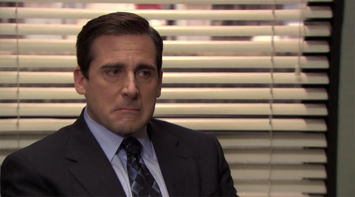 Michael Scott looking upset, wearing a suit and tie, in an office setting with blinds in the background.