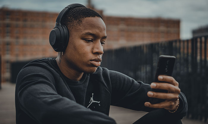 Young man wearing headphones, sitting outdoors and focused on his phone while enjoying music facts content.