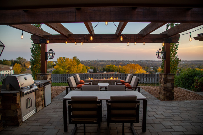 Outdoor pergola in an open backyard with seating area, fire pit, and grill overlooking a scenic autumn landscape.
