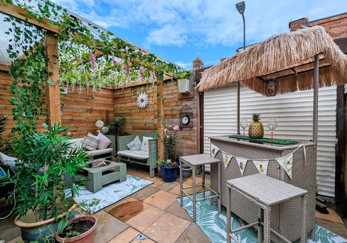 Backyard pergola decorated with flowers and greenery, featuring cozy seating and a tiki-style bar under a vibrant blue sky.