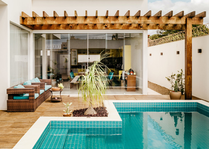 Backyard poolside pergola with wooden beams, wicker seating, and turquoise cushions overlooking a modern swimming pool.