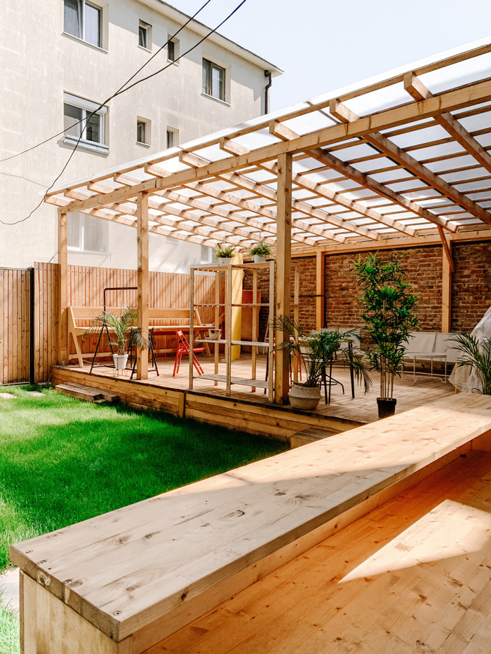 Wooden pergola with a see-through roof on a patio surrounded by green grass and outdoor plants in a backyard setting.
