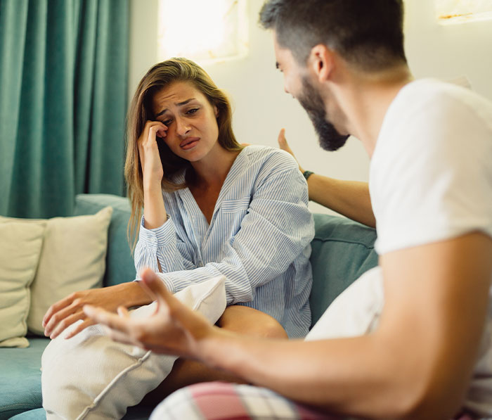 Upset woman and her fianc&eacute; sitting on couch, discussing family rejection and revealing his earnings in a tense moment.