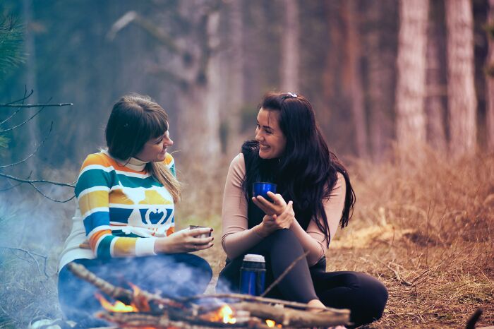 Two women sitting by a campfire in a forest, sharing deep conversations in an online group setting.