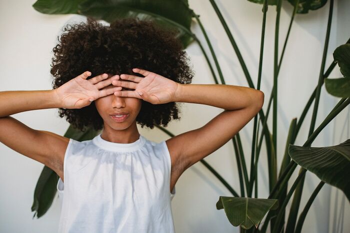 Person with curly hair covering eyes with hands, surrounded by large green leaves, pondering what disappeared unnoticed.