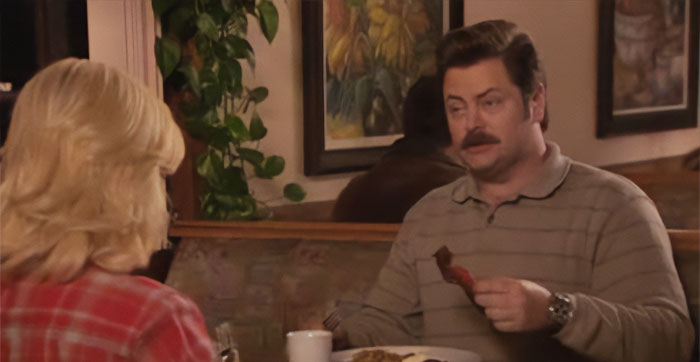 Ron Swanson holding and eating a piece of bacon while talking in a cozy diner setting, illustrating Ron Swanson quotes.