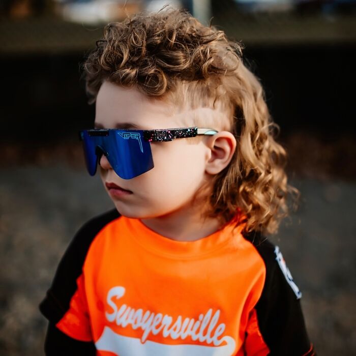 Young boy with a curly mullet hairstyle wearing blue sunglasses and an orange shirt outdoors, showcasing the cheddar wiz mullet.
