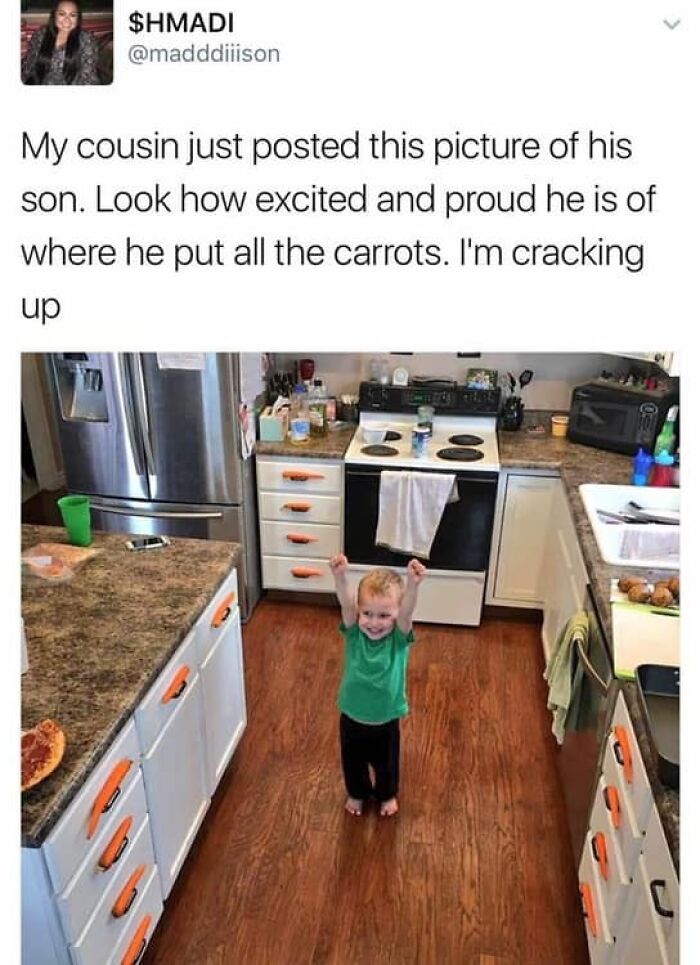Toddler proudly poses in kitchen next to drawers labeled with carrots; funny clean pic for a good laugh.