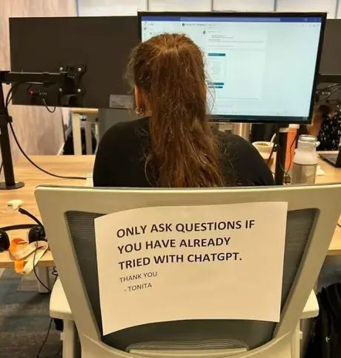 Office worker's chair with a sign about using ChatGPT before asking questions at a computer desk.