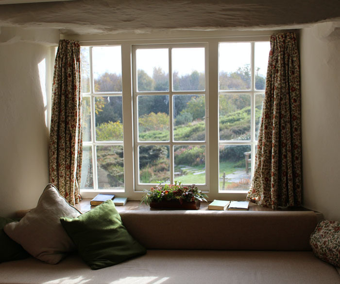 Cozy room with patterned curtains and cushions overlooking a tall hedge outside a countryside window.