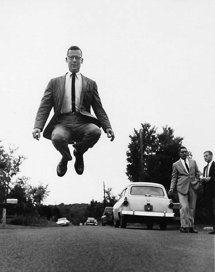 Man in a suit jumping mid-air on a road with vintage cars and two men walking in the background historical photos.