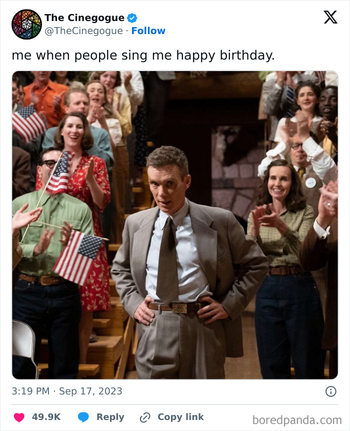 Man in a suit surrounded by cheering crowd holding flags, looking unimpressed during a birthday celebration.