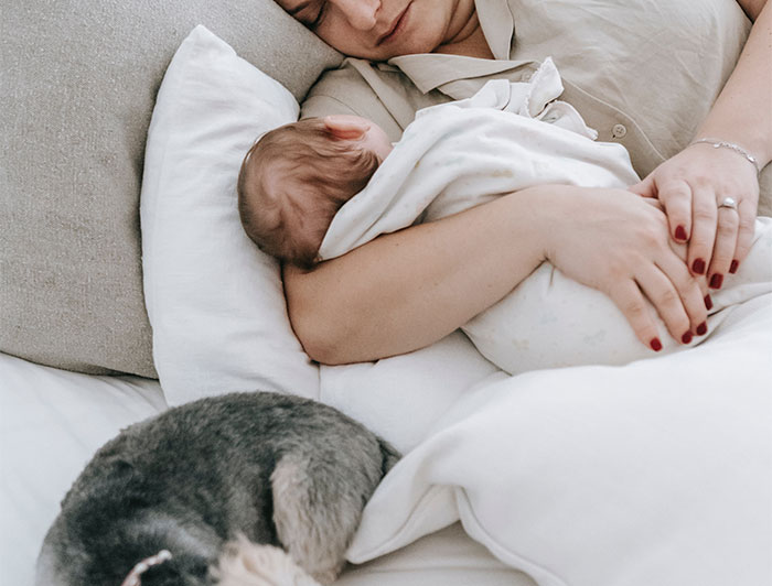 Woman resting on bed holding baby wrapped in blanket with a dog lying nearby, symbolizing woman’s marriage and sacrifice. Woman resting on bed holding baby wrapped in blanket with a dog lying nearby, symbolizing woman’s marriage and sacrifice.