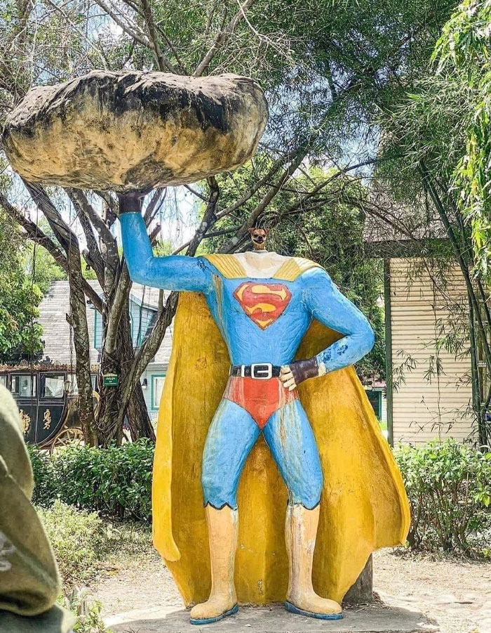 A dog meme featuring a statue of Superman with a large rock above his head. However, instead of Superman's head, the statue has a dog's head, creating a humorous and surreal image. The setting appears to be an outdoor area with trees and buildings in the background. The text above the image is not included, focusing solely on the funny juxtaposition of the dog's head on the superhero's body.