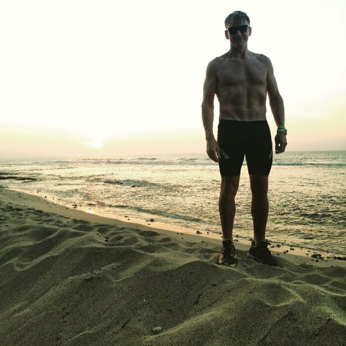 Shirtless man with visible muscles standing on the beach during sunset, showcasing his really buff physique.