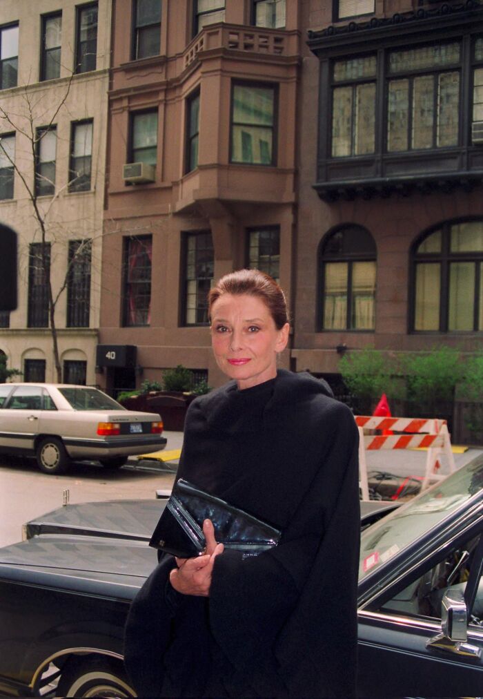 Audrey Hepburn wearing a black outfit holding a clutch purse on a city street in a historical photo.
