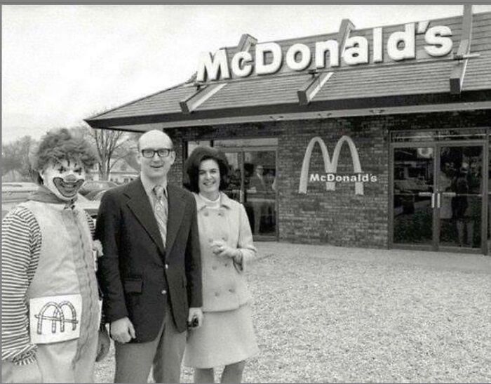 Vintage historical photo of three people, including a clown and a couple, standing outside an early McDonald's restaurant.