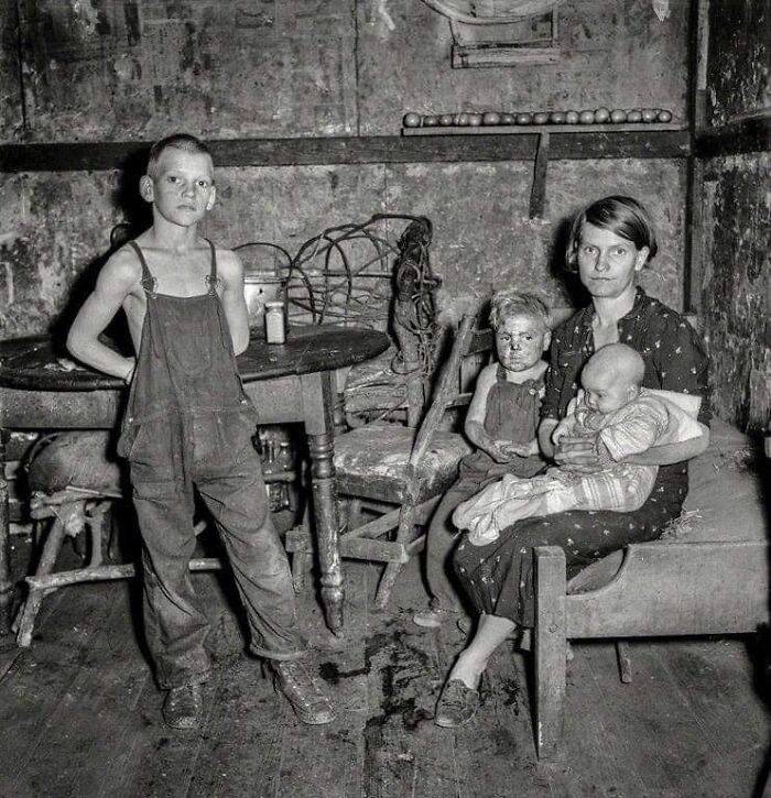 Black and white historical photo of a woman with three children in a worn, rustic interior setting.