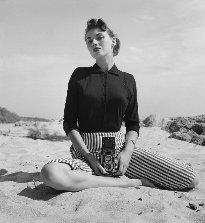 Young woman sitting on the beach holding a vintage camera in this historical photo from the past.