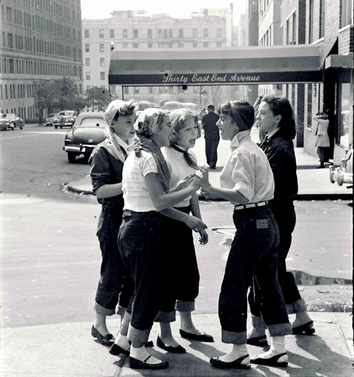 Group of young women dressed in 1950s style clothing socializing on a city street in a historical photo.