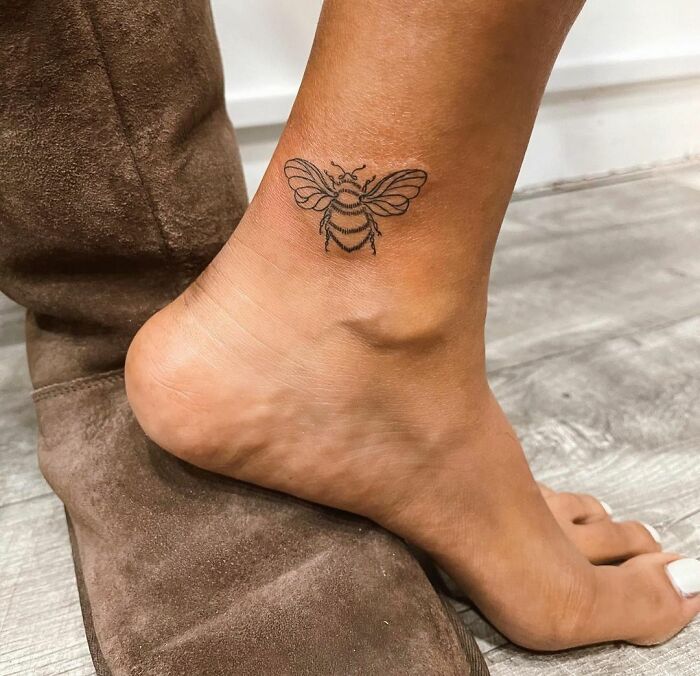 Bee ankle tattoo design inspiration on skin next to brown suede shoe.