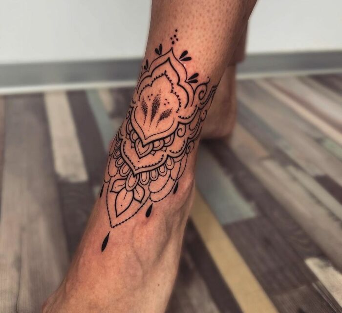 Intricate ankle tattoo with mandala design on a wooden floor backdrop.