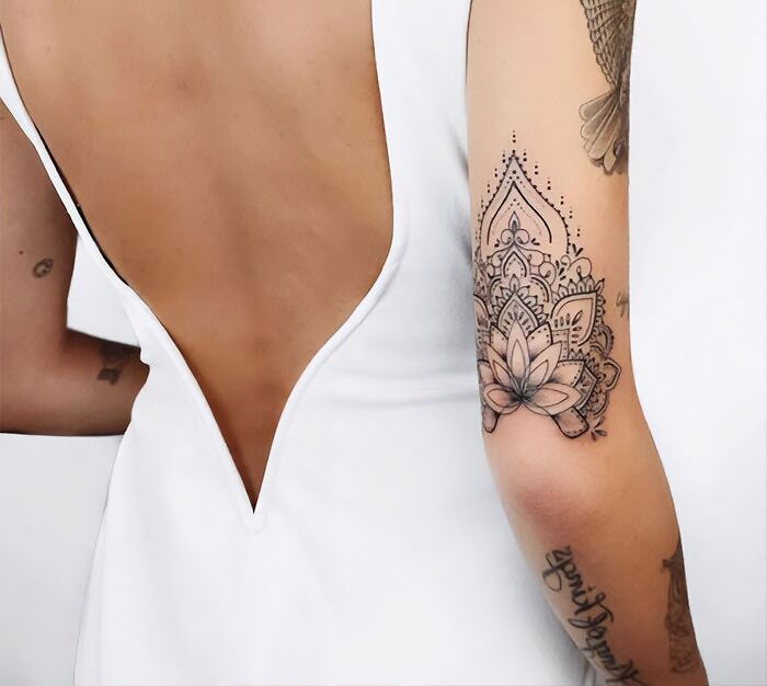 Intricate mandala elbow tattoo on person's arm wearing a white dress, showcasing detailed linework and floral design.