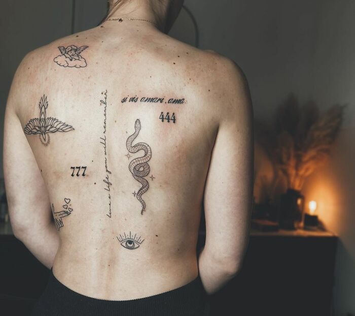 Person's back with unique patchwork tattoos including a snake, numbers, and symbolic designs in a dimly lit room.