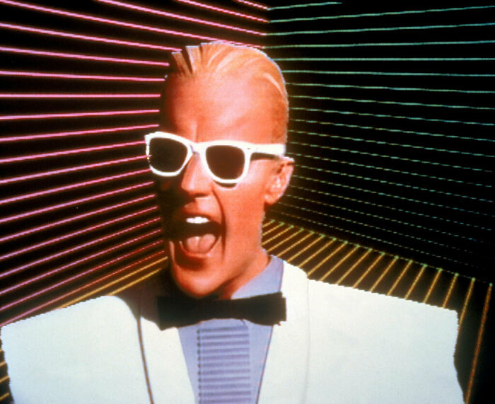 Man with slicked hair and sunglasses, wearing a white suit, set against a retro striped background, embodying a futuristic theme.